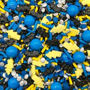 May include: A close-up of a mix of colorful sprinkles. The sprinkles include blue spheres, blue rods, yellow lightning bolts, black bat shapes, and silver nonpareils. These sprinkles are likely for decorating baked goods.