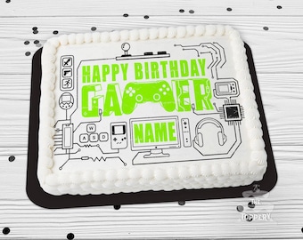 May include: A rectangular white cake with a black base, decorated with a green and black gamer-themed design. The cake features the text "HAPPY BIRTHDAY GAMER" and a space for a name, along with gaming icons.