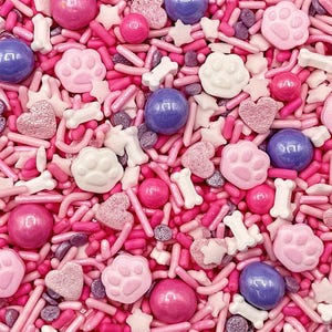 May include: A close-up of a mix of pink, white, and purple sprinkles, candies, and decorative shapes. The mix includes pink and white paw prints, bone shapes, hearts, and round candies. The background is filled with pink sprinkles.