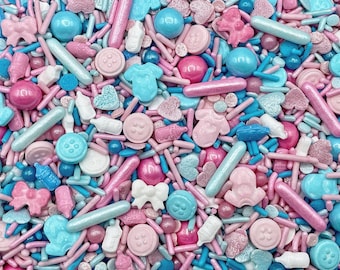 May include: A collection of edible sprinkles in shades of pink and blue, featuring various shapes including small baby bottles, onesies, bows, buttons, hearts, and spheres, mixed with thin rod-shaped sprinkles in a dense, colorful assortment.