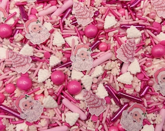May include: A festive mix of edible cake decorations featuring pink and white sugar sprinkles, shiny metallic pink rods, round pink gumballs, small white snowflake shapes, white Christmas tree shapes, and flat Santa Claus face sugar toppers.