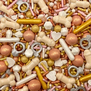 May include: A variety of sprinkles in gold, silver, pink, and white. The sprinkles are shaped like rings, hearts, bones, and other geometric shapes.