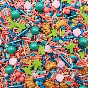 May include: A colorful mix of edible sprinkles featuring small pink seashells, green palm trees, gold crowns, teal round sugar pearls, metallic blue rods, and thin rod-shaped sprinkles in shades of pink, blue, and orange for cake decoration.