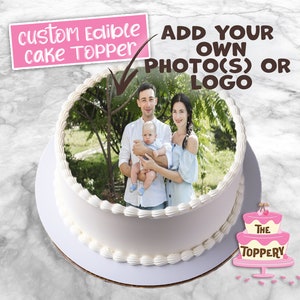 May include: A round white cake with a family photo edible cake topper. The cake has a decorative border and text that reads "custom Edible cake Topper" and "ADD YOUR OWN PHOTO(S) OR LOGO". The cake also has a logo that reads "The Toppery".