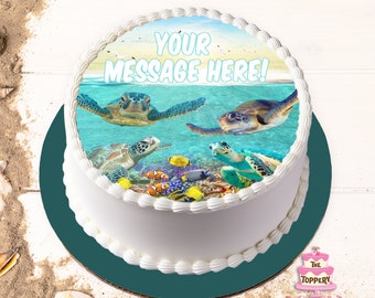 May include: A white cake with a round edible image of sea turtles swimming in a blue ocean with coral reefs and colourful fish. The text "YOUR MESSAGE HERE!" is printed on the image.