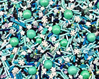 May include: A collection of edible cake sprinkles featuring a mix of light blue, turquoise, and dark blue rod-shaped jimmies, white star and snowflake shapes, small black sugar pearls, and large smooth mint green round candy balls.