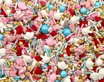 May include: A colorful mix of edible cake sprinkles featuring white rabbit shapes, pink flamingo figures, light blue spheres, red hearts, and gold metallic beads, combined with thin pink and light blue rod-shaped sprinkles for decoration.