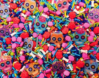 May include: A colorful mix of edible sprinkles featuring flat sugar skulls in various shades of blue, orange, pink, and purple, interspersed with small red rose-shaped candies, white bone-shaped pieces, and thin rod-shaped sprinkles.