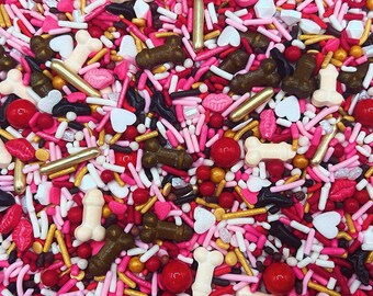 May include: A colourful mix of edible cake sprinkles featuring pink, red, white, and gold hues. The assortment includes rod-shaped jimmies, round sugar pearls, heart-shaped pieces, lip-shaped candies, and phallic-shaped sugar decorations.