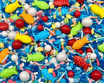 May include: A colourful mix of edible sprinkles featuring small fish shapes in yellow, orange, green, and blue, alongside red round sweets, white pearl-like spheres, tiny yellow stars, and blue rod-shaped sprinkles scattered across the surface.