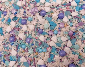 May include: A festive mix of edible sprinkles featuring white snowflake shapes, light blue and purple round candy balls, small white Christmas tree shapes, and thin rod-shaped sprinkles in shades of blue, purple, and white for cake decoration.