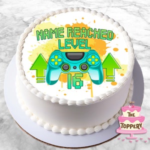 May include: A white cake with a green and blue video game controller, yellow splatters, and the text "NAME REACHED LEVEL 16" on the top.