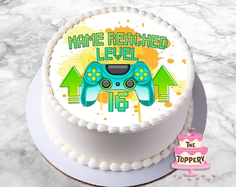 May include: A white cake with a green and blue video game controller, yellow splatters, and the text "NAME REACHED LEVEL 16" on the top.