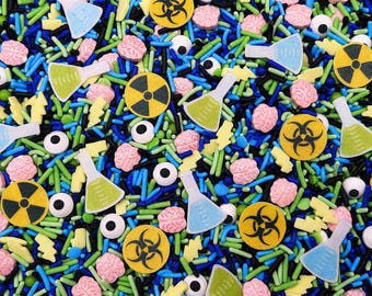 May include: A colorful mix of edible sprinkles featuring yellow biohazard symbols, pink brain shapes, white eyeballs with black pupils, and various laboratory flasks filled with blue or green liquid, interspersed with blue, green, and black rods.