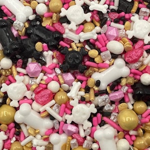 May include: A mix of edible cake sprinkles featuring white and black bone shapes, white and black skull and crossbones, metallic gold spheres, pink sugar crystals, and small pink and white rod-shaped sprinkles for decorative baking.