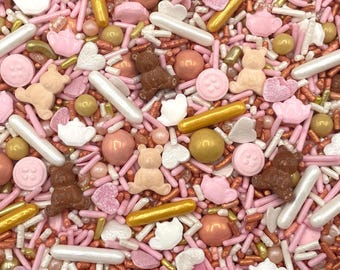 May include: A decorative mix of edible sprinkles featuring light pink, white, and metallic gold colors. The assortment includes small teddy bear shapes, round buttons, hearts, tiny teapots, long rod-shaped sprinkles, and round metallic balls.