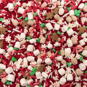May include: A festive mix of edible sprinkles featuring red and white candy canes, brown gingerbread men, white snowflake shapes, green Christmas trees, and small white cylindrical marshmallows scattered across a dense, colorful surface.