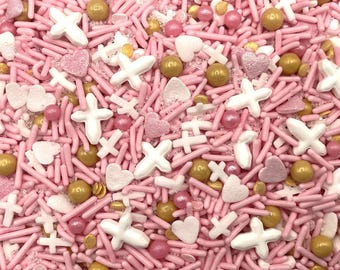 May include: A decorative mix of edible sprinkles featuring light pink rod-shaped jimmies, white cross-shaped pieces, white heart-shaped sugar shapes, metallic gold spherical dragees, and small pink pearlized balls scattered across the surface.
