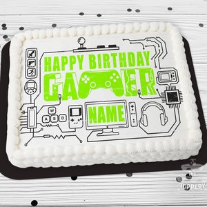 May include: A white cake with green frosting and black icing. The cake is decorated with a gamer theme, including a joystick, a game controller, and the words "Happy Birthday Gamer".