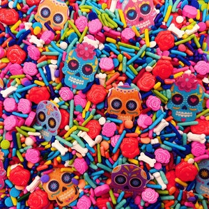 May include: A colorful mix of sprinkles and candy shaped like skulls and flowers. The sprinkles are in various colors, including red, blue, green, yellow, and pink. The candy is in the shape of skulls with different colored designs and flowers with white centers and pink petals.