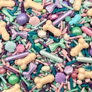 May include: A colorful mix of edible sprinkles featuring light peach bone shapes, iridescent purple mermaid tails, pastel pink seashells, and light blue seahorses, interspersed with various shades of purple, green, and pink rod and ball sprinkles.