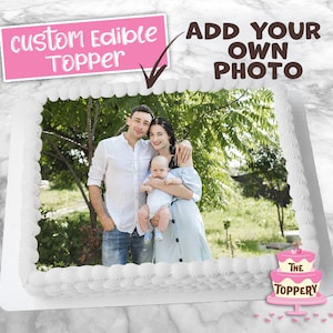 May include: A white cake with a custom edible photo topper. The photo shows a family of three, a couple and their baby, standing in a park. The cake is decorated with a white frosting border and the words "The Toppery" are printed on the bottom right corner.