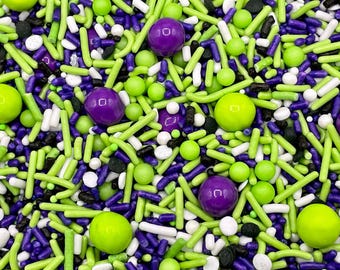 May include: A collection of edible cake sprinkles featuring a mix of lime green, purple, white, and black shapes. The assortment includes long rod-shaped sprinkles, small round nonpareils, and larger round candy balls in a vibrant colour palette.