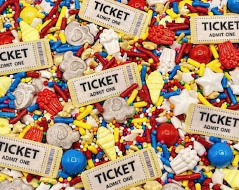 May include: A colorful mix of edible cake sprinkles featuring rectangular beige tickets with the text TICKET ADMIT ONE, red and blue candy spheres, yellow ice cream cone shapes, white stars, and various small red, blue, and yellow rod-shaped sprinkles.