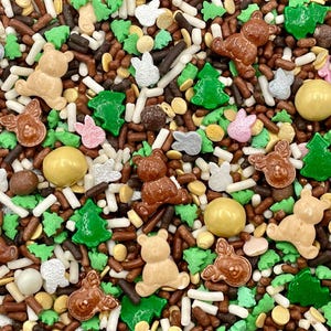 May include: A festive mix of edible sprinkles featuring brown teddy bear shapes, green Christmas tree silhouettes, light pink and silver bunny shapes, brown and gold round candy balls, and a variety of brown, white, and green rod-shaped sprinkles.