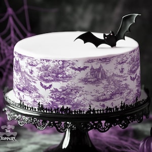 May include: A round cake with a white top and a purple toile-style pattern featuring haunted houses, trees, and bats around the sides. A black bat-shaped topper is placed on the top surface. The cake sits on a black decorative metal cake stand.