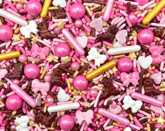 May include: A collection of decorative sprinkles featuring pink, brown, white, and gold elements. The mix includes small rod-shaped sprinkles, round spheres, white and pink bows, brown cowboy boots, and small white and pink hearts.