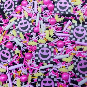 May include: A colorful mix of sprinkles and decorative pieces in pink, white, black, and yellow. The mix includes small round sprinkles, rod-shaped sprinkles, and square pieces with a smiling face and heart eyes.