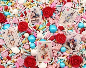 May include: A mix of edible sprinkles featuring rectangular playing cards with Alice in Wonderland illustrations, red rose shapes, light blue spheres, pink hearts, white rabbit silhouettes, and gold rods. The cards depict characters like the White Rabbit.