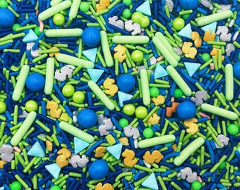May include: A colorful mix of edible sprinkles featuring dark blue, light green, and bright blue rod-shaped pieces, round spheres, light blue triangles, and metallic silver and gold dollar sign shapes scattered across the entire surface.