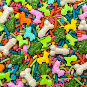 May include: A colorful assortment of edible sprinkles featuring white phallic shapes, green cacti, pink llamas, yellow llamas, orange llamas, and turquoise llamas mixed with blue, orange, and green rod-shaped sprinkles and small round confetti pieces.