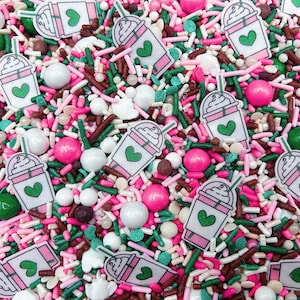 May include: A close-up of a variety of colorful sprinkles, including pink, green, brown, and white. The sprinkles are shaped like hearts, stars, and other fun designs. There are also small round candies in the mix.