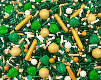 May include: A mix of edible sprinkles featuring dark green and light green rod-shaped jimmies, white and gold round sugar pearls, long gold rods, and bright green leaf-shaped sugar pieces scattered across a flat surface.