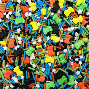 May include: A colorful mix of edible sprinkles featuring small game controller shapes in orange, green, yellow, and black, interspersed with blue, orange, and green rod-shaped sprinkles, small white star shapes, and tiny green round sugar balls.