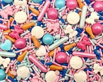 May include: A colorful mix of edible cake sprinkles featuring pink, blue, and white bows, round baseball-shaped candies, metallic pink and silver rods, orange bat-shaped pieces, and small blue, pink, and white spheres for decorative baking.