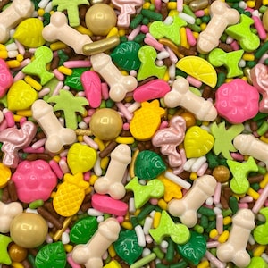 May include: A colorful assortment of edible cake sprinkles featuring various shapes including light beige phallic figures, pink flamingos, yellow pineapples, green palm leaves, bright pink flowers, and gold metallic spheres mixed with small rod-shaped sprinkles.