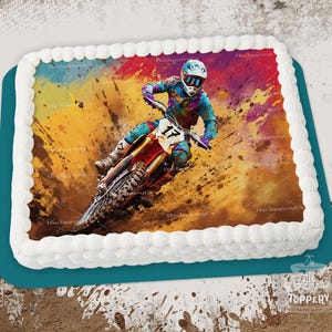 May include: A rectangular cake with white frosting and a colorful image of a motocross rider on a dirt bike. The rider wears a helmet and colorful gear. The cake is on a teal-colored base.