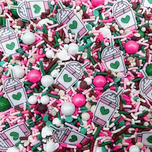 May include: A colorful mix of edible sprinkles featuring white, pink, and green rod-shaped sprinkles, round white and pink sugar pearls, and flat, white-and-pink iced coffee cup shapes with a green heart graphic printed on each cup.