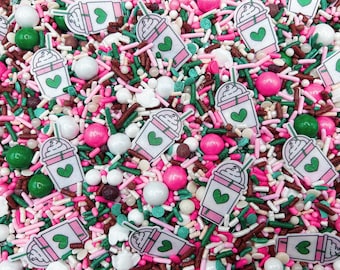 May include: A colorful mix of edible sprinkles featuring white, pink, and green rod-shaped sprinkles, round white and pink sugar pearls, and flat, white-and-pink iced coffee cup shapes with a green heart graphic printed on each cup.