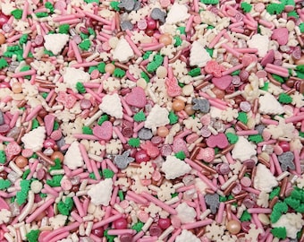 May include: A festive mix of edible sprinkles featuring pink, green, white, and metallic copper elements. The assortment includes small rod-shaped jimmies, round sugar pearls, flat heart shapes, snowflake cutouts, and small Christmas tree shapes.