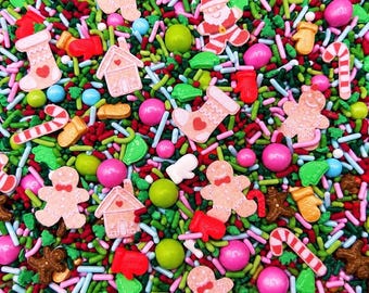 May include: A festive mix of edible sprinkles featuring gingerbread people, Santa Claus figures, gingerbread houses, stockings, candy canes, and mittens. The blend includes small rod-shaped sprinkles in red, green, and white, alongside round pink and green candies.