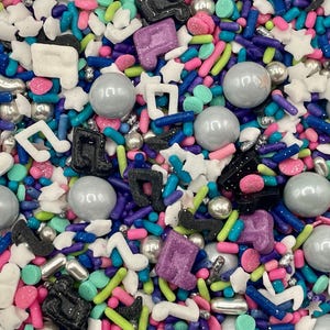 May include: A colorful mix of edible sprinkles featuring white, black, and purple musical note shapes, silver metallic spheres, small white stars, and thin rod-shaped sprinkles in shades of blue, pink, green, and purple for cake decoration.