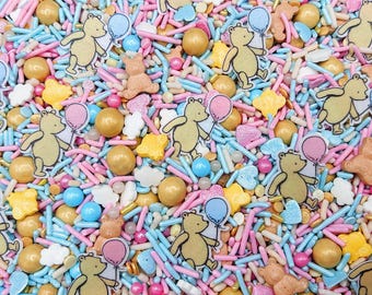 May include: A decorative sprinkle mix featuring edible yellow bear-shaped figures holding pink or blue balloons, interspersed with gold metallic spheres, small white cloud shapes, and a variety of pink, blue, and white rod-shaped sprinkles.