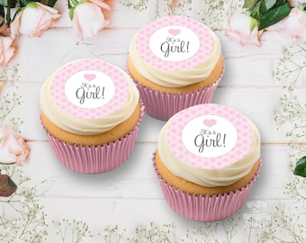 May include: Three cupcakes with pink wrappers and white frosting. Each cupcake has a pink and white circular topper with the text "It's a Girl!" and a heart. The cupcakes are arranged on a white wooden surface with floral decorations.