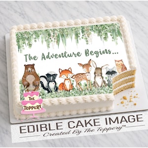 May include: A rectangular sheet cake with white frosting borders, topped with an edible image featuring watercolor forest animals including a bear, skunk, fox, deer, rabbit, raccoon, and owl, with the text The Adventure Begins in green.
