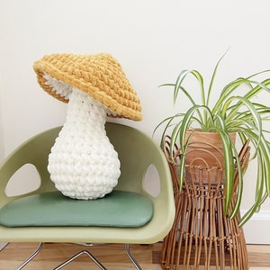 May include: A crocheted mushroom pillow with a white stem and a yellow cap. The pillow is sitting on a green chair with a wicker plant stand next to it.